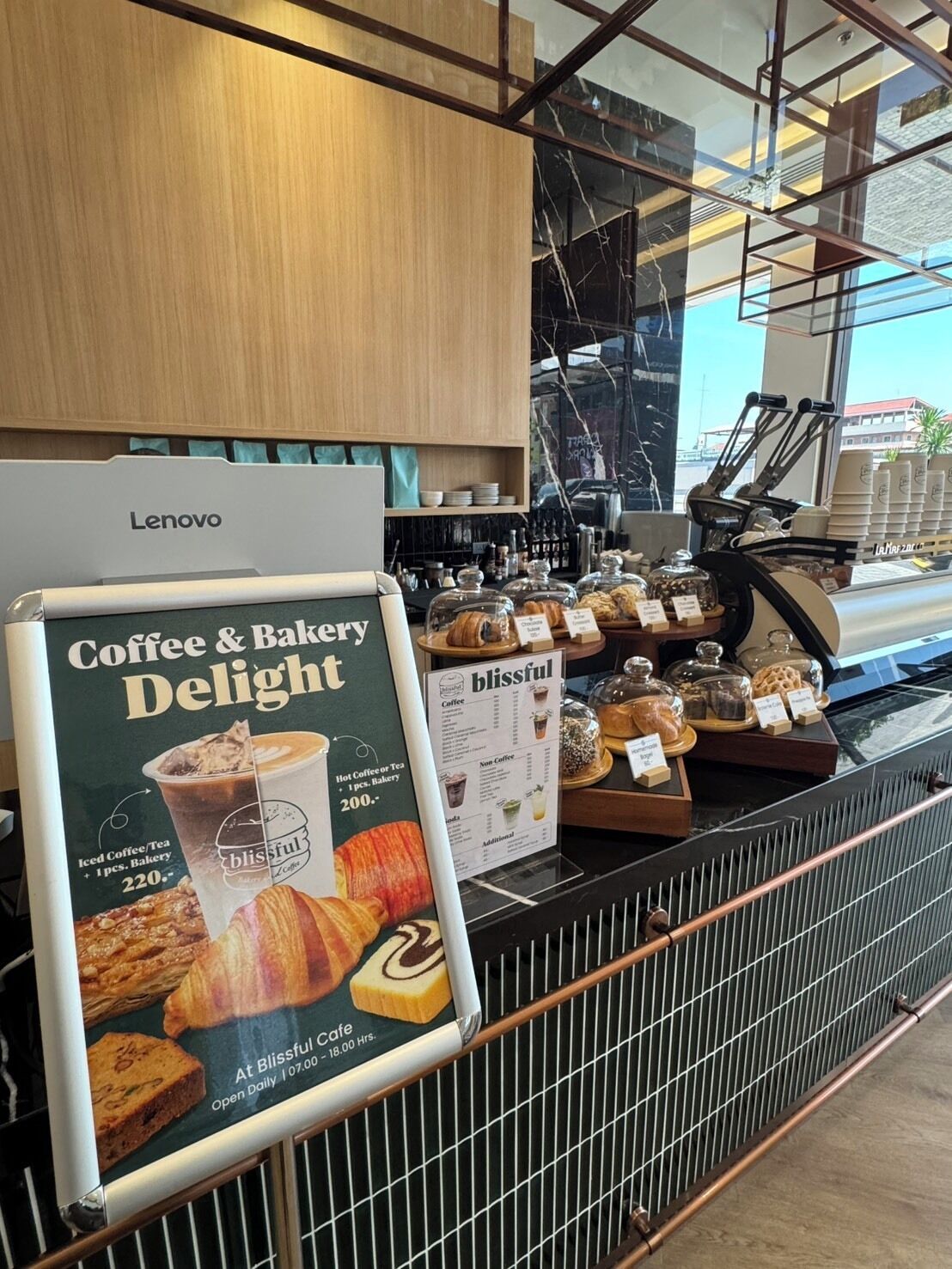 Mostrador de cafetería con una pizarra de menú, una computadora Lenovo y diversos productos de panadería a la vista.