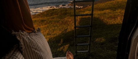 Una cama con almohadas a rayas, vista al océano a través de una ventana y una escalera apoyada en el marco.