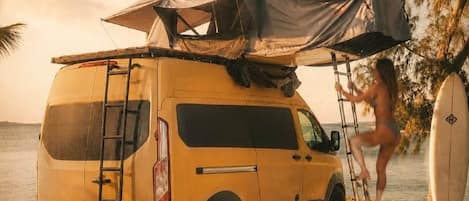 A yellow van parked on a sandy beach with a canopy, a surfboard, and a ladder.