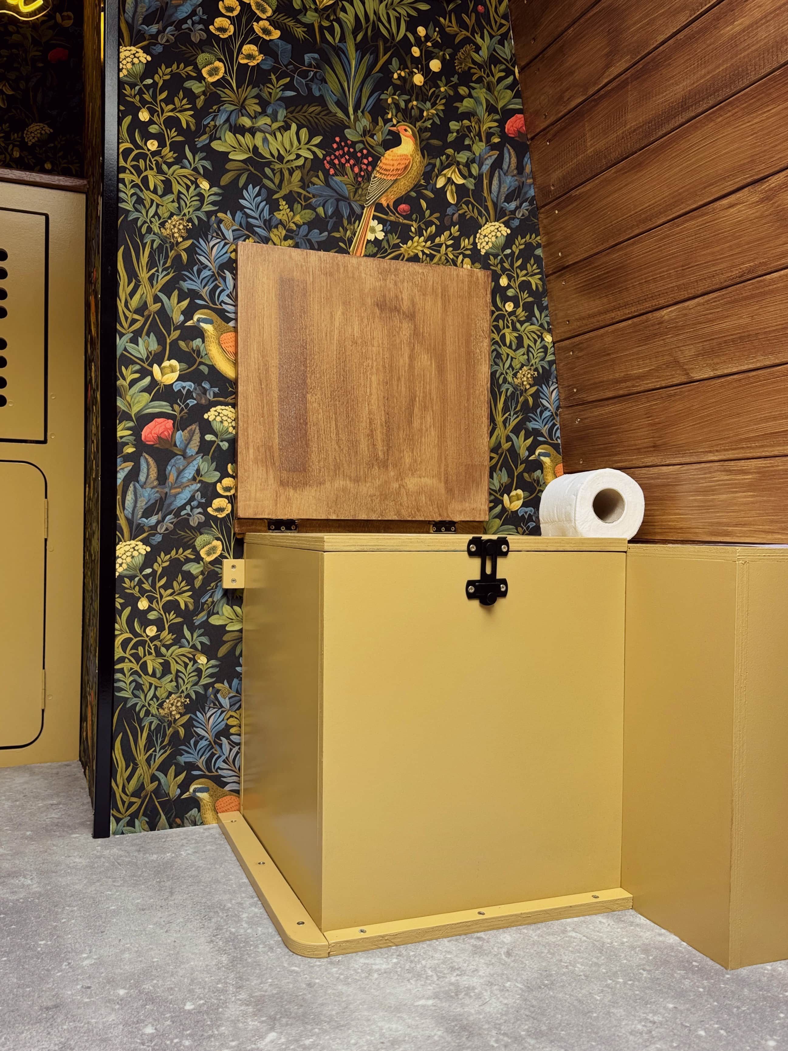 Un meuble jaune avec une boîte en bois posée dessus, un rouleau de papier toilette et un papier peint à motifs floraux.