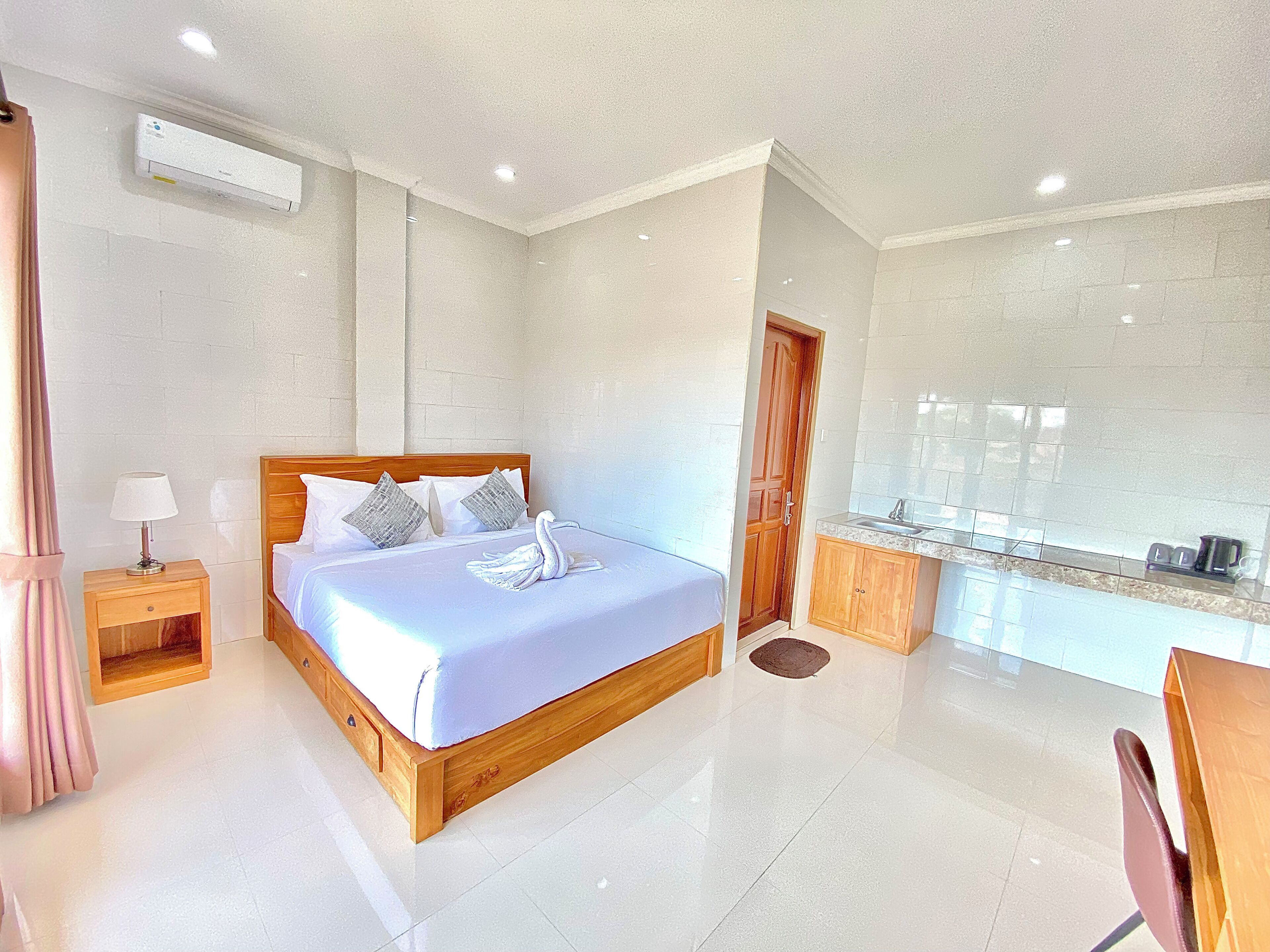 A hotel room with a bed, a nightstand, a lamp, a swan decoration, a wooden door, a kitchen area with a sink, and a chair.