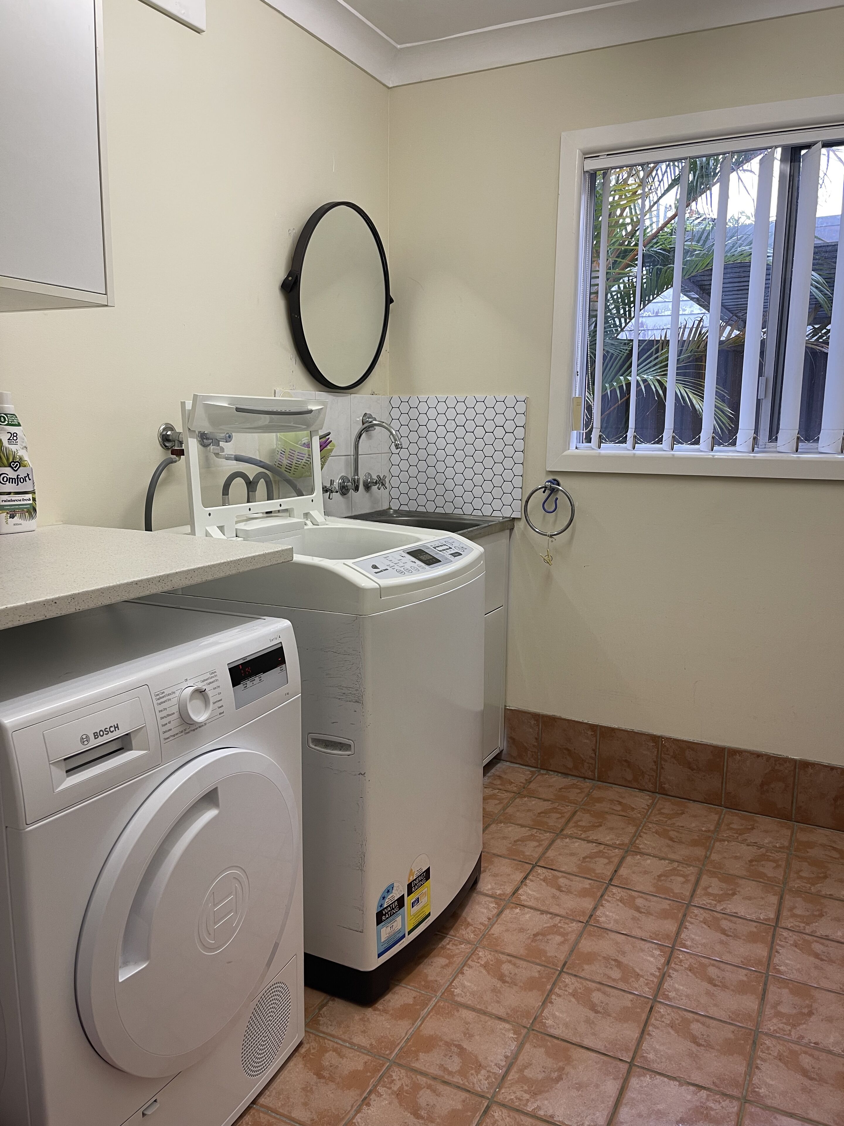 Un espace buanderie équipé d’un lave-linge Bosch, d’un sèche-linge et d’une fenêtre donnant sur des palmiers.
