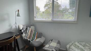 Ein Zimmer mit Schreibtisch, einem gemusterten Sessel, einem kleinen Hocker, einem Bett mit blattbasiertem Bezug und einem Fenster mit Jalousien.