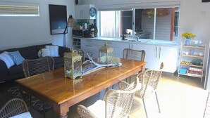 A dining area with a wooden table and wicker chairs, a kitchen with white cabinets, and a window with blinds.