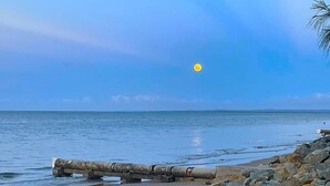 A beach with a calm sea, a clear sky, and a sun in the distance.