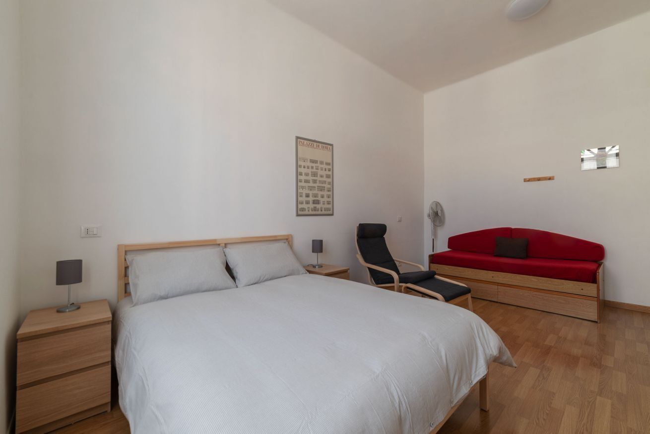 A bedroom with a bed, a nightstand, a chair, a red sofa, and a wall-mounted fan.