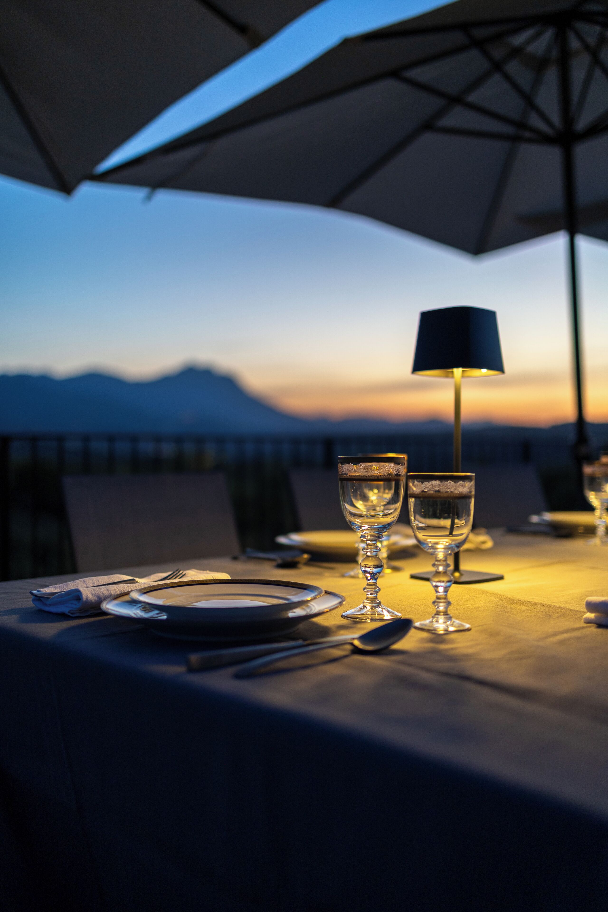 A table set for two with a lamp, glasses, and cutlery, overlooking a mountainous landscape.
