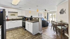 A modern kitchen with white cabinets, a central island, and a dining area with a glass table and wicker chairs. - Seas You Later - 2 Bedroom Condo Just 2 Blocks from the Beach with Large Pool, Elevator (Oak Island)