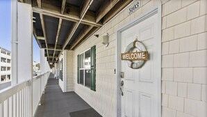 A hotel corridor with a welcome sign, a door with a number, and a view of the outside buildings. - Seas You Later - 2 Bedroom Condo Just 2 Blocks from the Beach with Large Pool, Elevator (Oak Island)