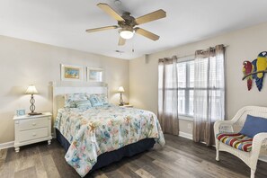 Une chambre à coucher comprenant un lit, des tables de chevet, une chaise, un ventilateur de plafond et une fenêtre avec des rideaux.