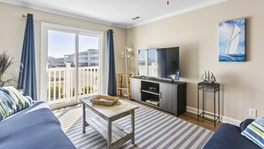 A living room with a sofa, a coffee table, a TV, and a balcony with curtains. - Seas You Later - 2 Bedroom Condo Just 2 Blocks from the Beach with Large Pool, Elevator (Oak Island)