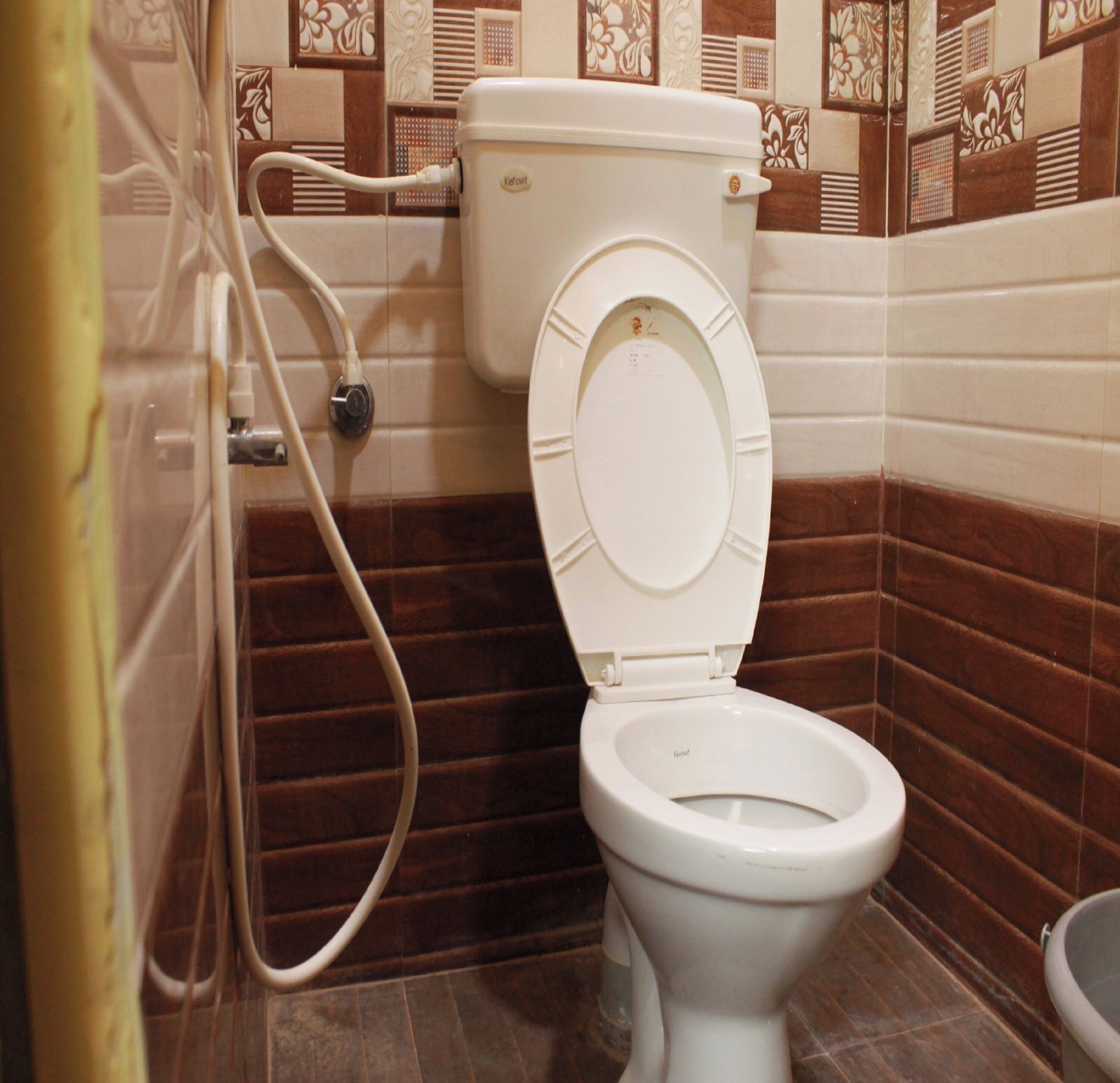 A white toilet with a tank and a bidet attachment in a bathroom with brown and beige patterned tiles.