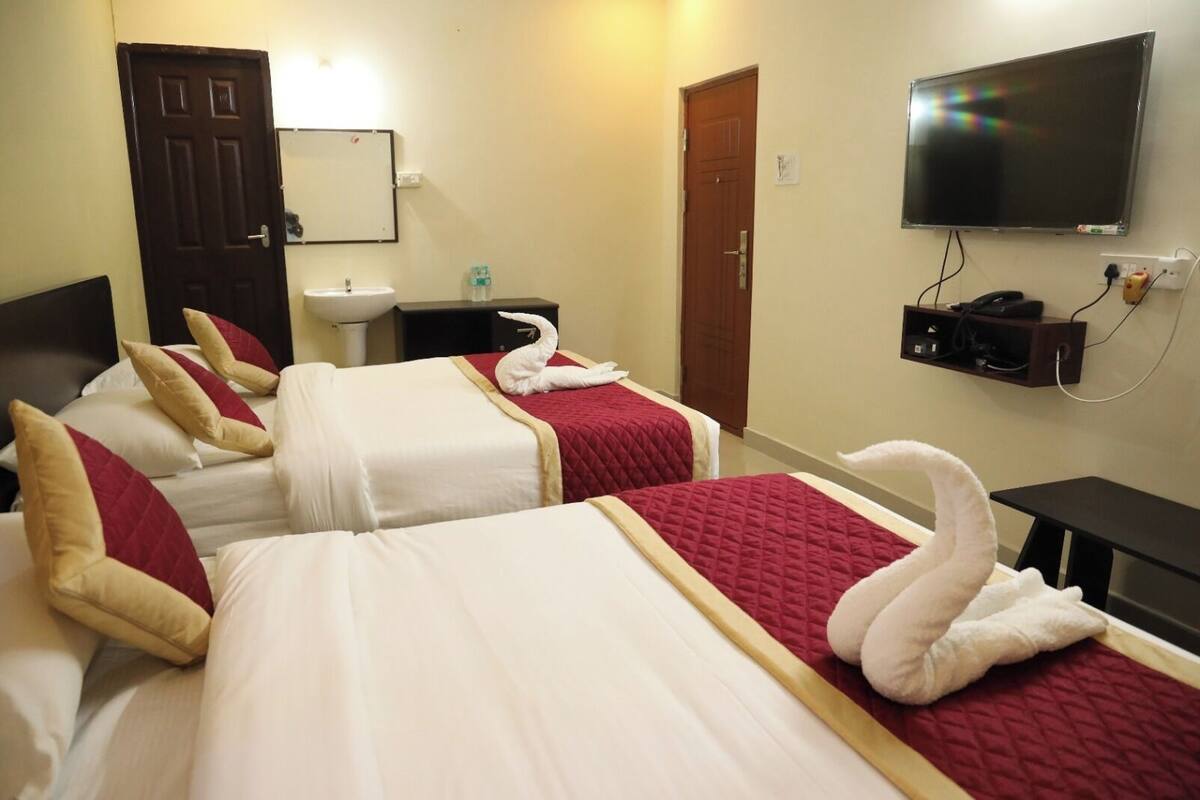 A hotel room with two beds, each with a red and white bedspread, and a white swan-shaped pillow. There is a TV mounted on the wall, a small table with a lamp, and a door in the background.