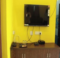 A television mounted on a yellow wall, connected to various electronic devices and a telephone on a wooden cabinet.