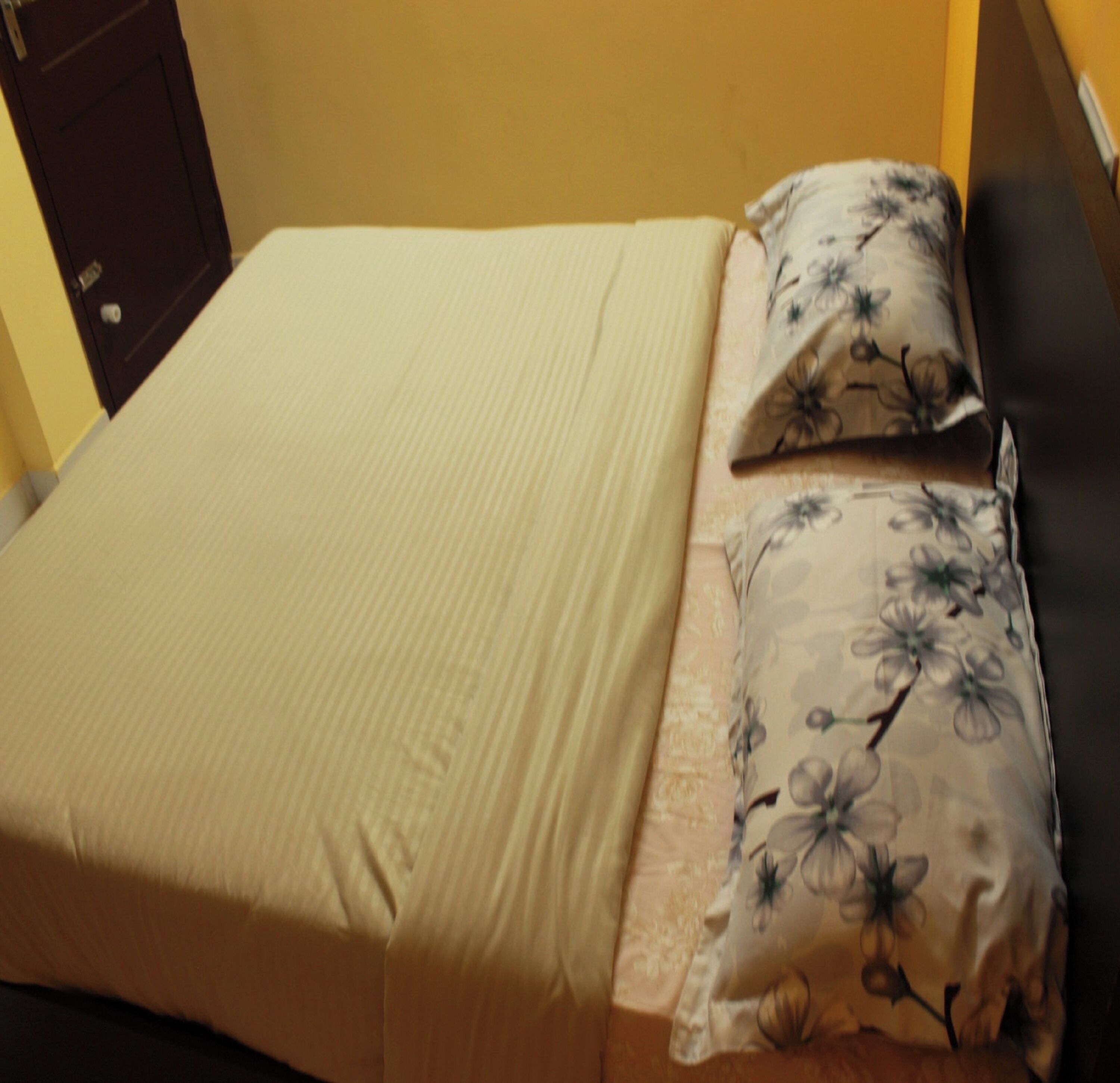 A bed with a floral pillow and a plain bedspread, a wooden nightstand, and a door in the background.
