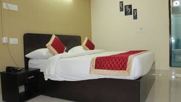 Una cama bien hecha con ropa blanca y detalles rojos, una mesita de noche con un teléfono, y una pared con un enchufe y un cuadro pequeño.
