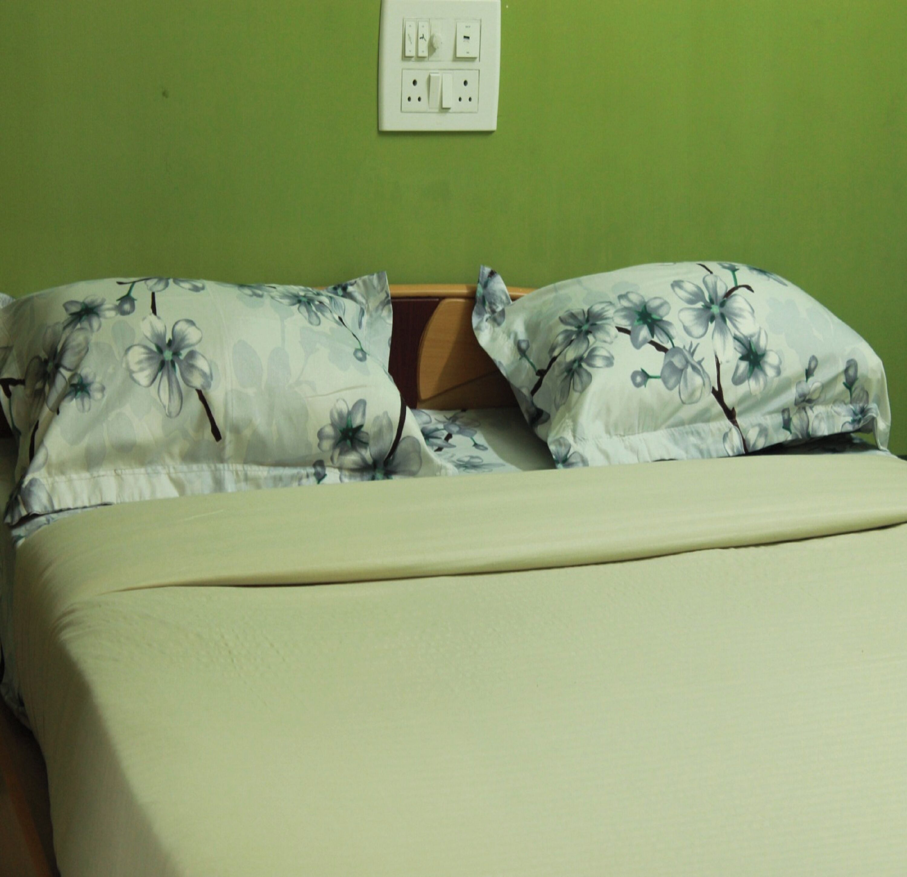 A bed with floral-patterned pillows and a plain bedspread in a room with a green wall and a white electrical outlet above the bed.