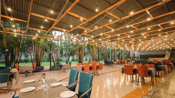 A spacious indoor dining area with tables and chairs arranged for guests, featuring a high ceiling with exposed beams and string lights, and a view of a lush garden outside. - Yatri Niwas By Hotel Suraj Inn (Silvassa)