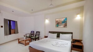 A hotel room with a large bed, two chairs, a small table, a painting on the wall, and a ceiling fan. - Yatri Niwas By Hotel Suraj Inn (Silvassa)