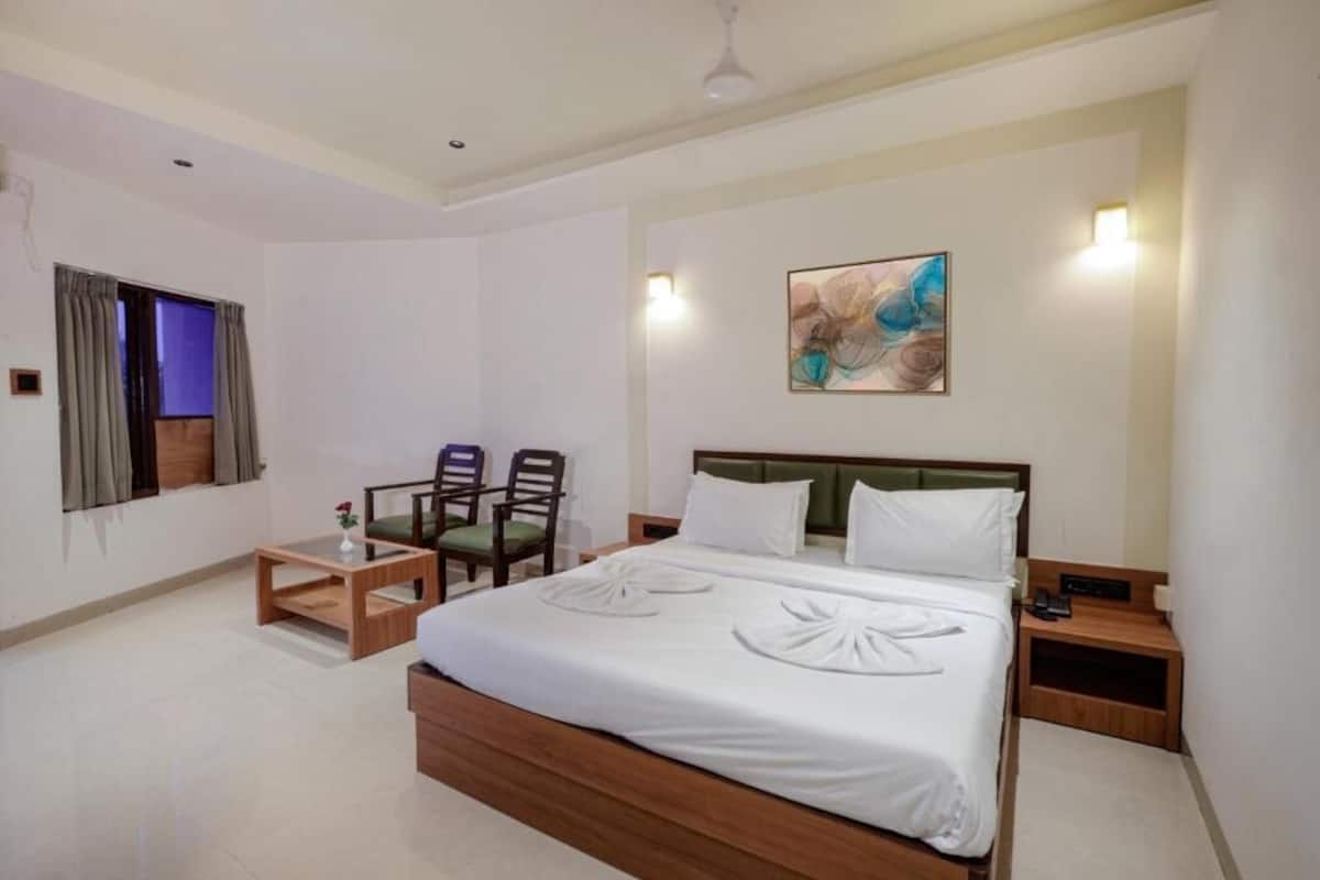 A hotel room with a large bed, two chairs, a small table, a painting on the wall, and a ceiling fan.