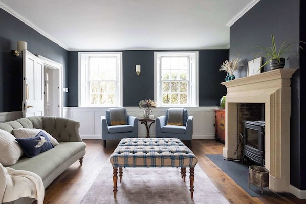 A room with a dark blue wall, a fireplace, a sofa, two armchairs, and a table with a vase of flowers.