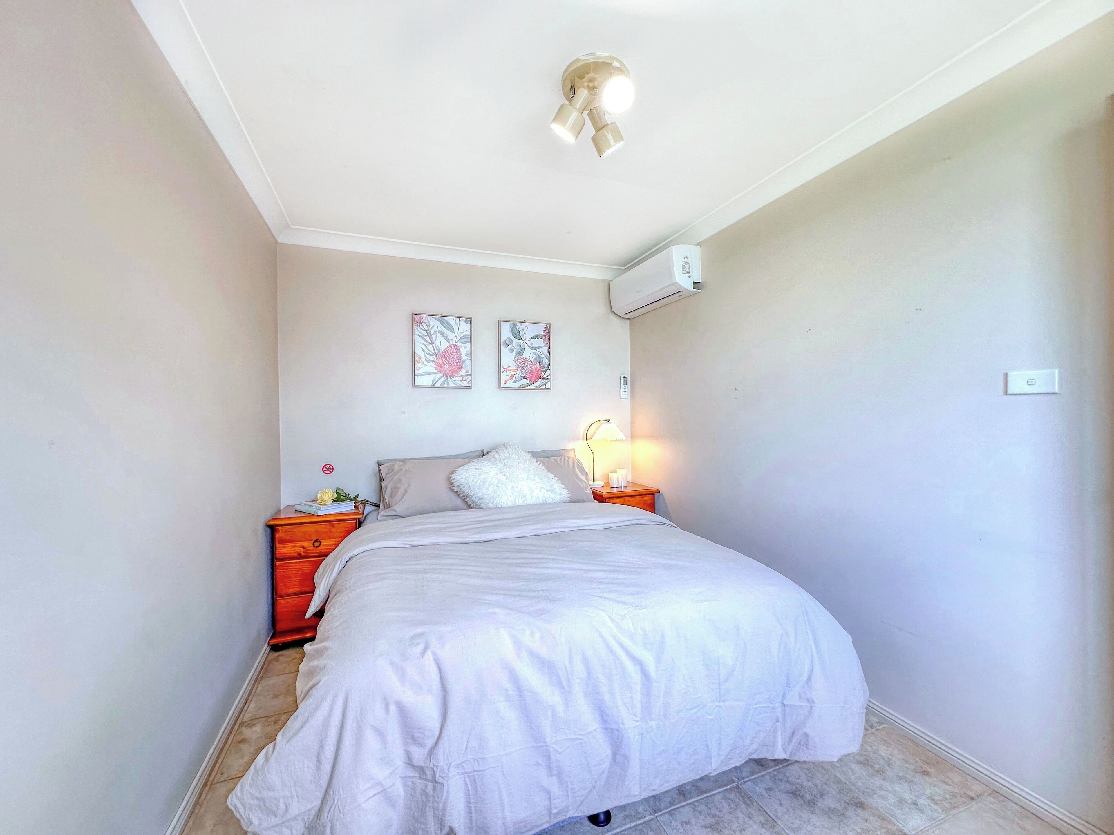 A bedroom with a bed, nightstands, a wall-mounted air conditioner, and two framed floral pictures.