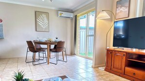 Un comedor con una mesa preparada para cuatro personas, un televisor sobre un mueble de madera y una planta en maceta sobre una mesa.