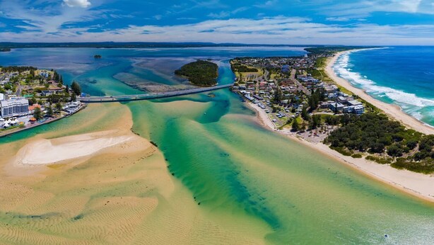 Vista aérea de un pueblo costero con un puente que conecta dos extensiones de tierra, aguas azules cristalinas y una playa de arena.