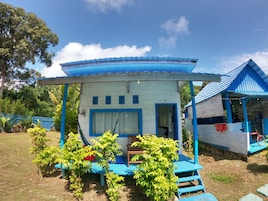 A small, single-story building with a blue roof and white walls, featuring a porch with steps and a bench.