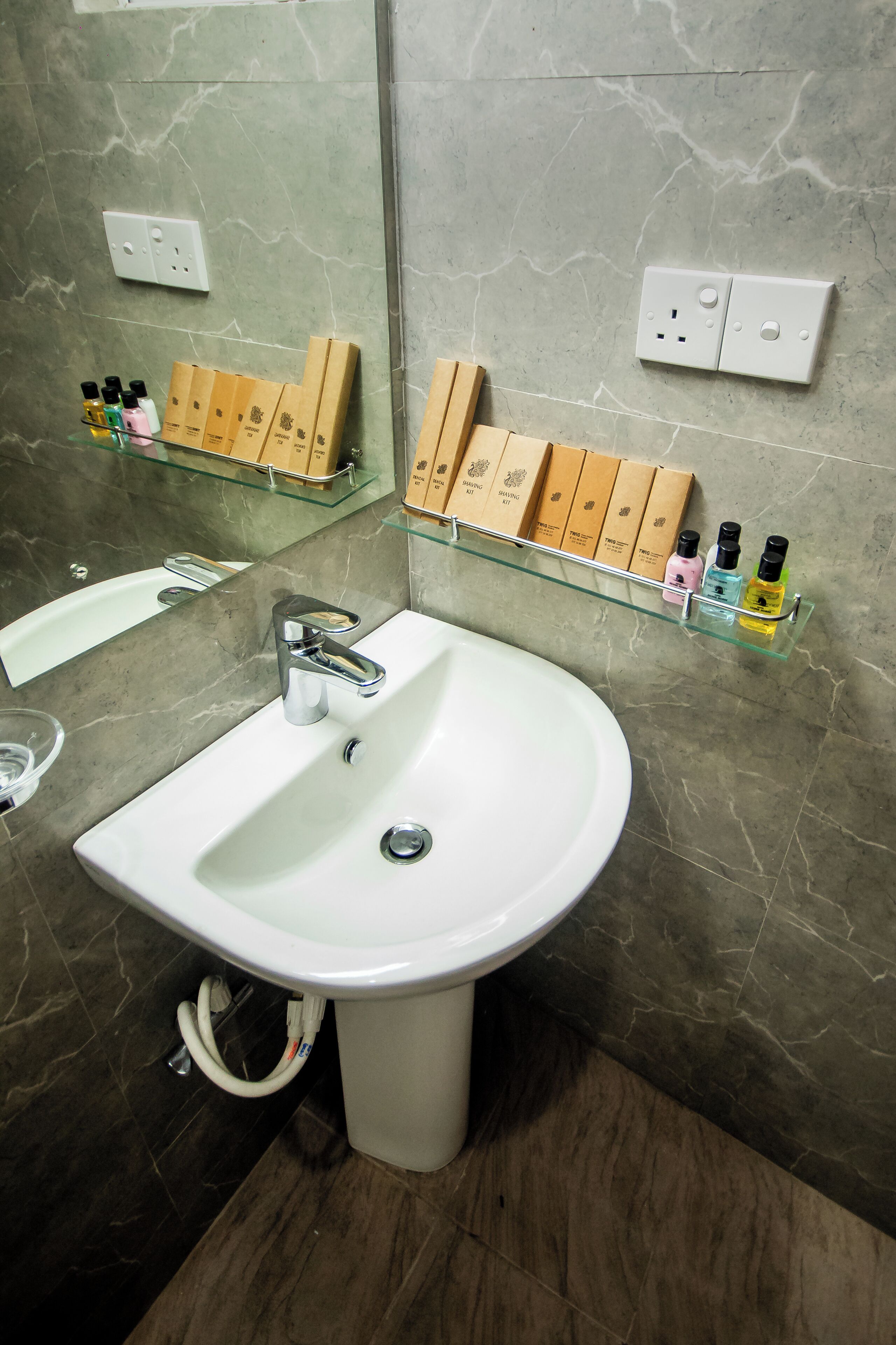 Une salle de bain avec un lavabo blanc, une étagère en verre où sont rangés les articles de toilette et un mur en marbre.