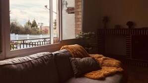 Un soggiorno con divano, camino e vista su una recinzione bianca e alberi.