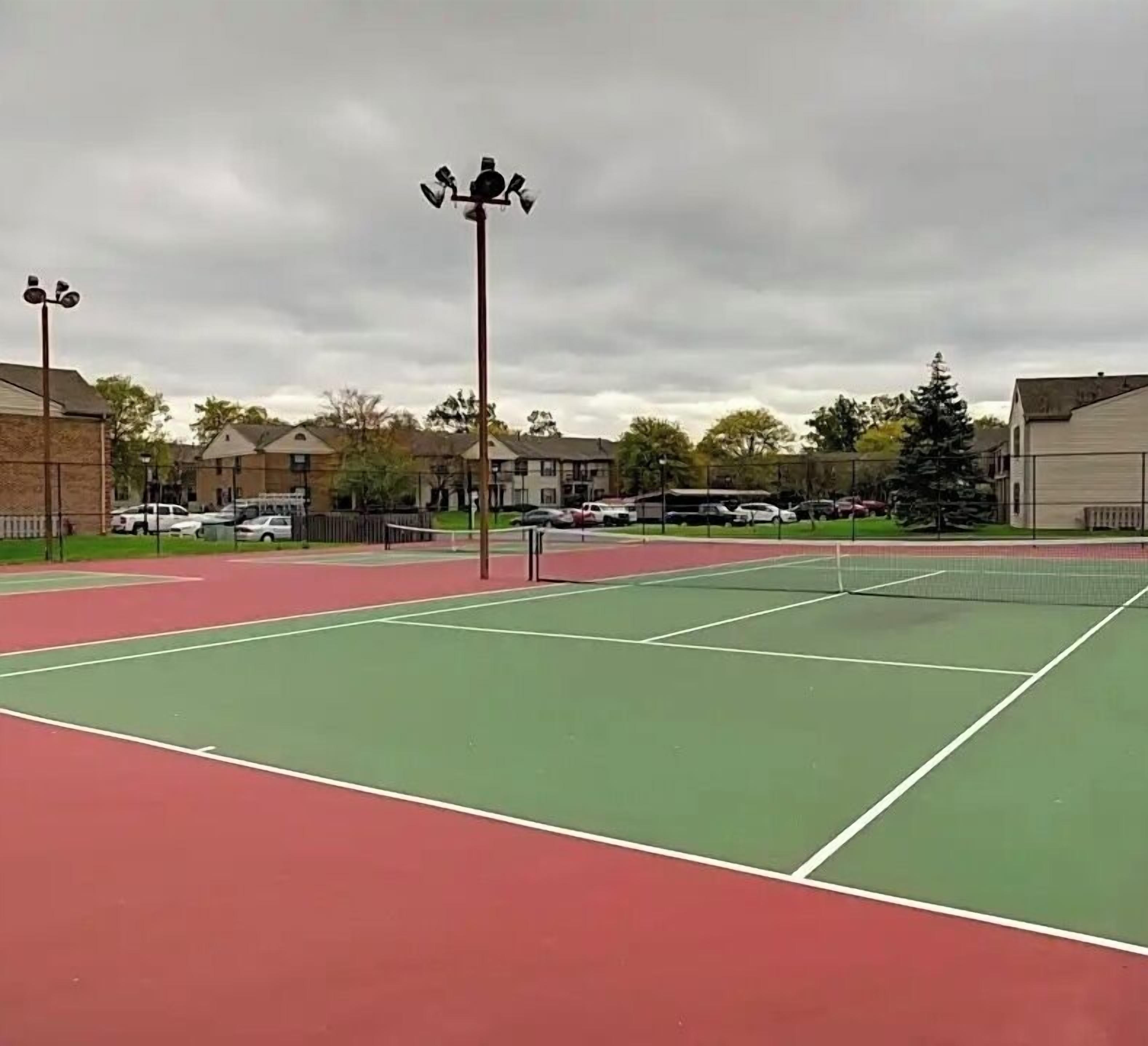 Ein Tennisplatz im Freien mit Netz, umgeben von einem Zaun und angrenzenden Gebäuden.