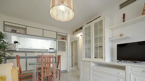 Una cocina moderna con armarios blancos, un comedor con sillas de madera, un televisor de pantalla plana grande y una lámpara colgante de mimbre.
