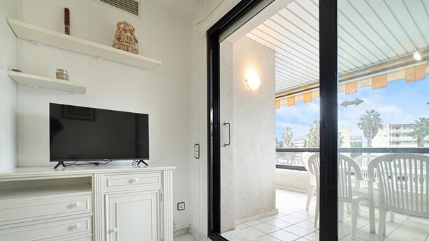 Una habitación con una televisión sobre un mueble blanco, un balcón con sillas y vistas a palmeras y edificios.