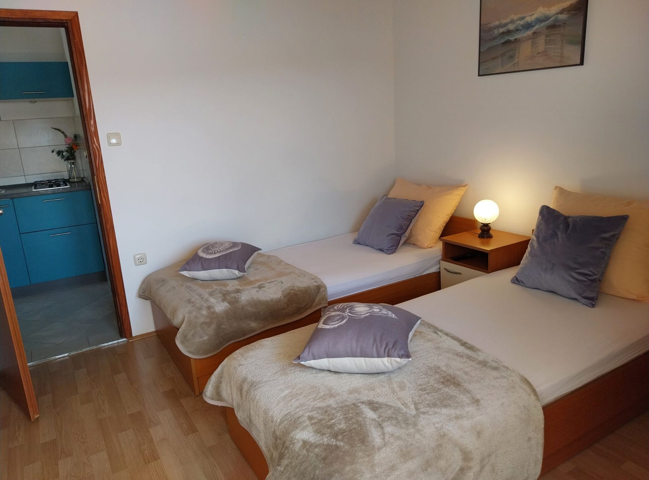 Een kleine, netjes ingerichte kamer met twee eenpersoonsbedden, een nachtkastje met een lamp, en uitzicht op een keukenhoek.
