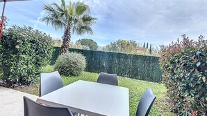 Une terrasse extérieure avec une table et des chaises, entourée de haies bien entretenues et d’un grand palmier.