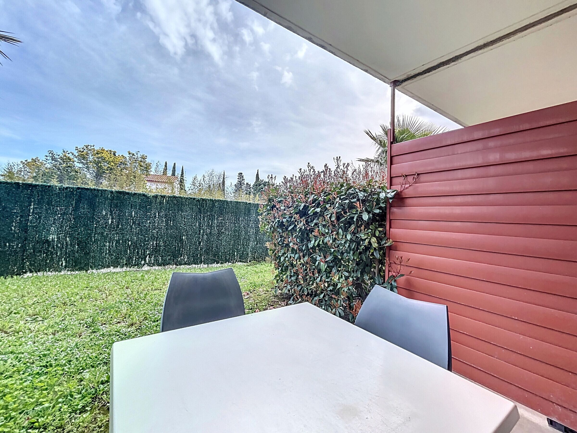 An outdoor patio area with a table and chairs, surrounded by a green hedge and a red wall.