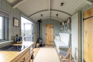 A cozy cabin interior with a wooden bench, a stove, and a window overlooking a forest. - Luxurious Shepherds Huts, Outdoor Baths, Sea Views (Devon)