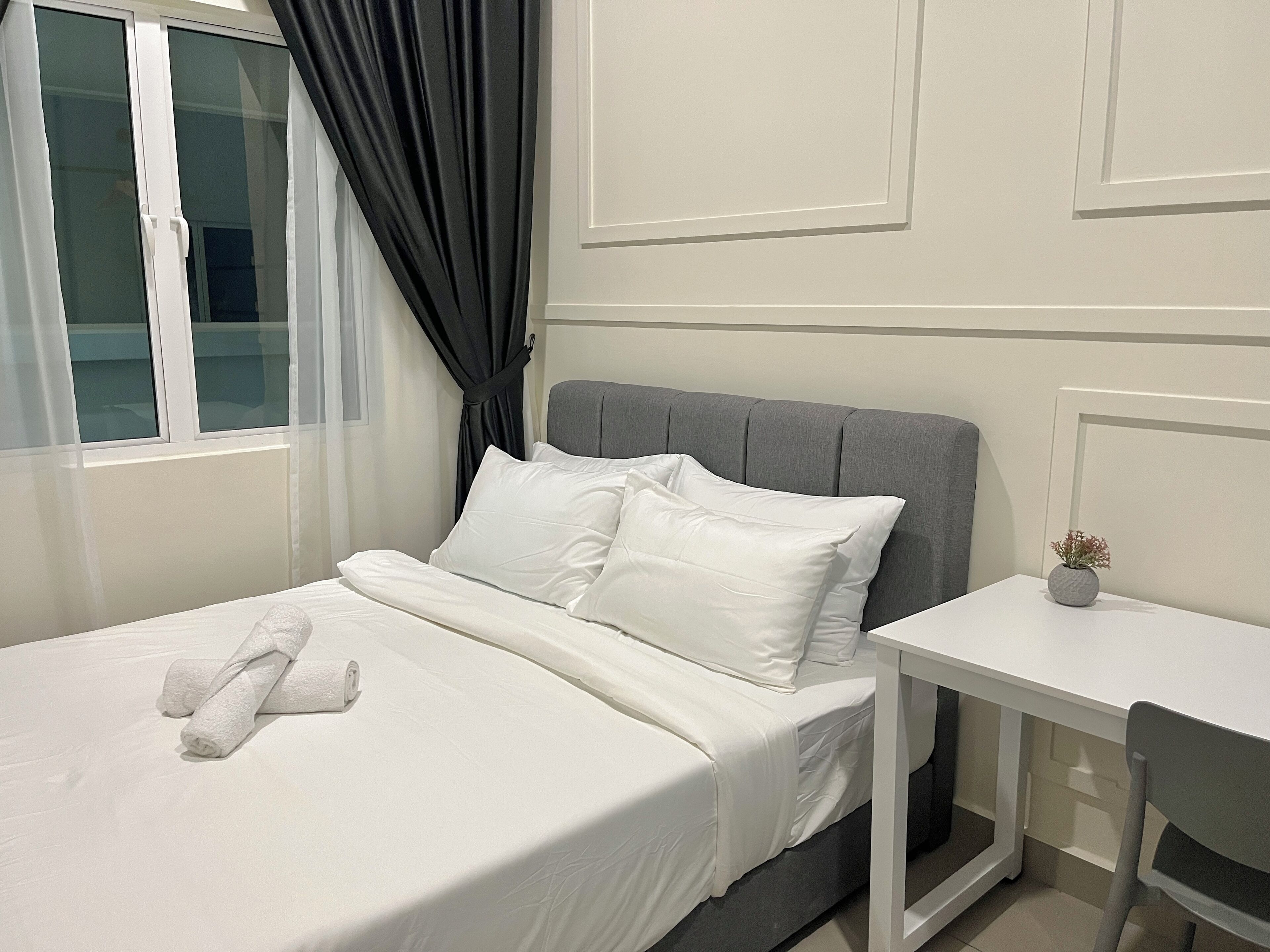Uma cama bem arrumada com lençóis e almofadas brancas, um painel de cabeceira estofado em cinza, uma pequena mesa de cabeceira com um vaso, e uma janela com cortinas brancas.