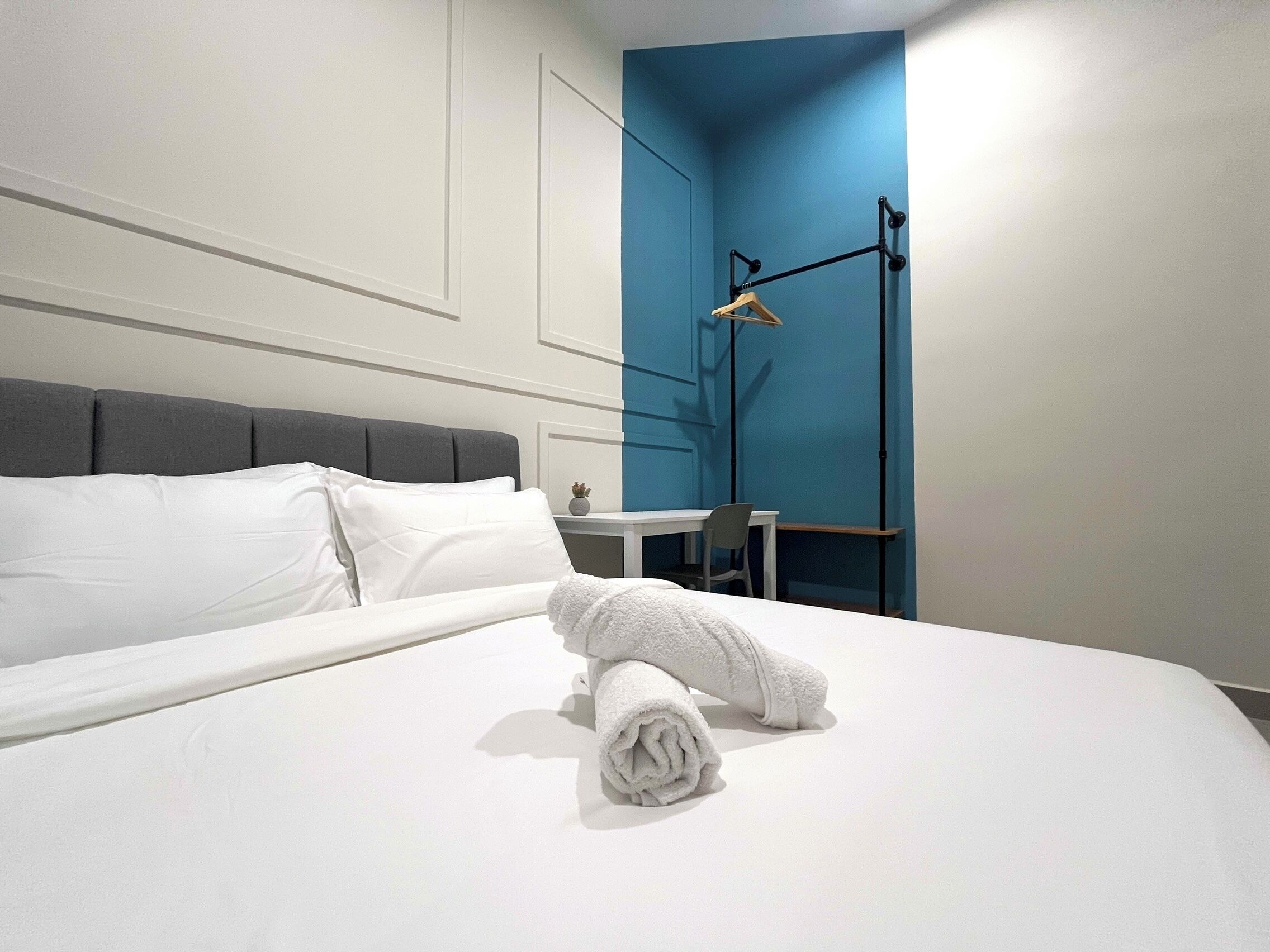 Un lit bien fait, avec du linge de lit blanc et une serviette enroulée, une tête de lit rembourrée de couleur grise, une petite table avec une chaise et une lampe sur pied.