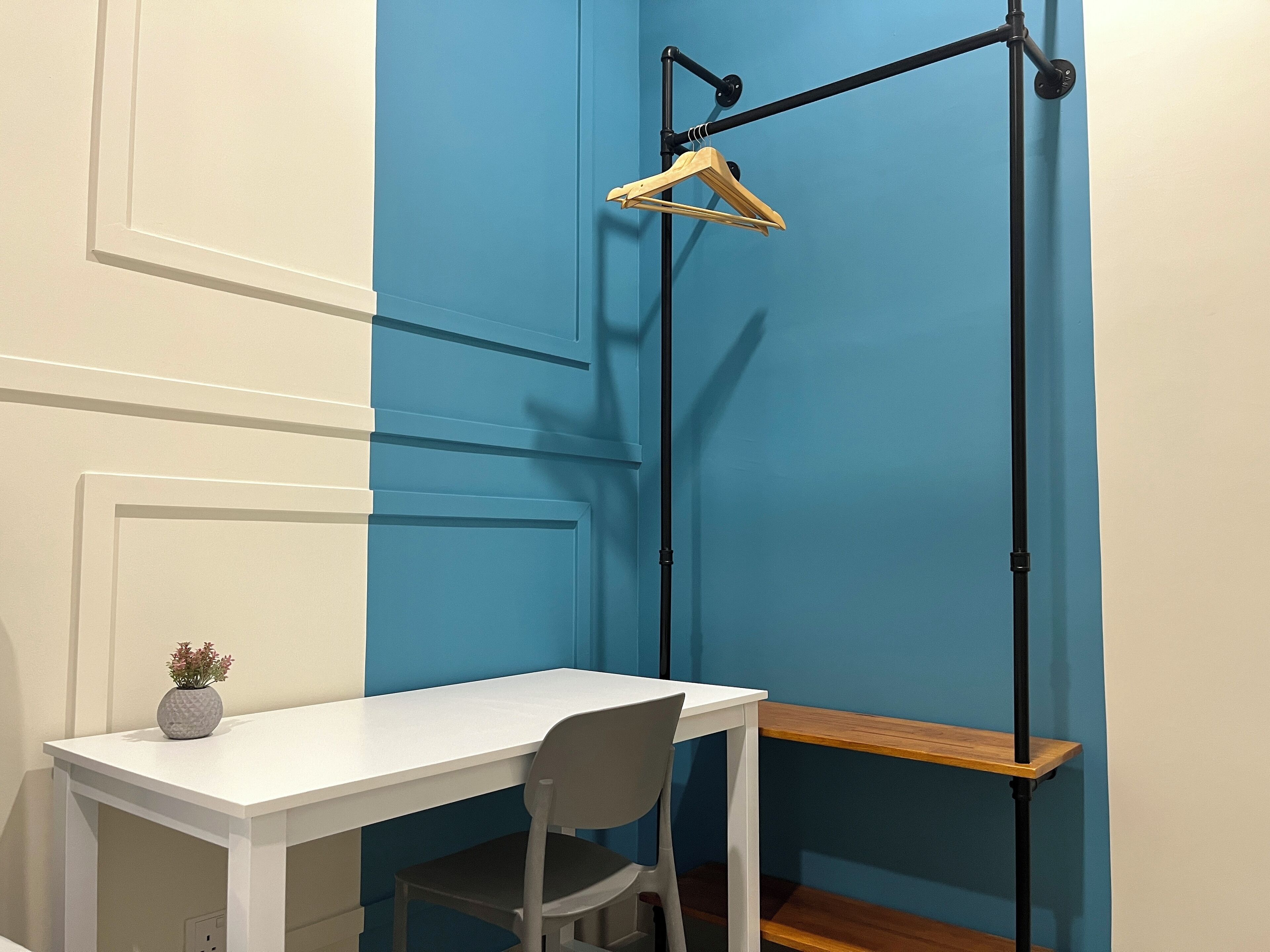 Una habitación con un escritorio blanco, una silla gris, un estante de madera y una barra colgante negra con un perchero de madera.