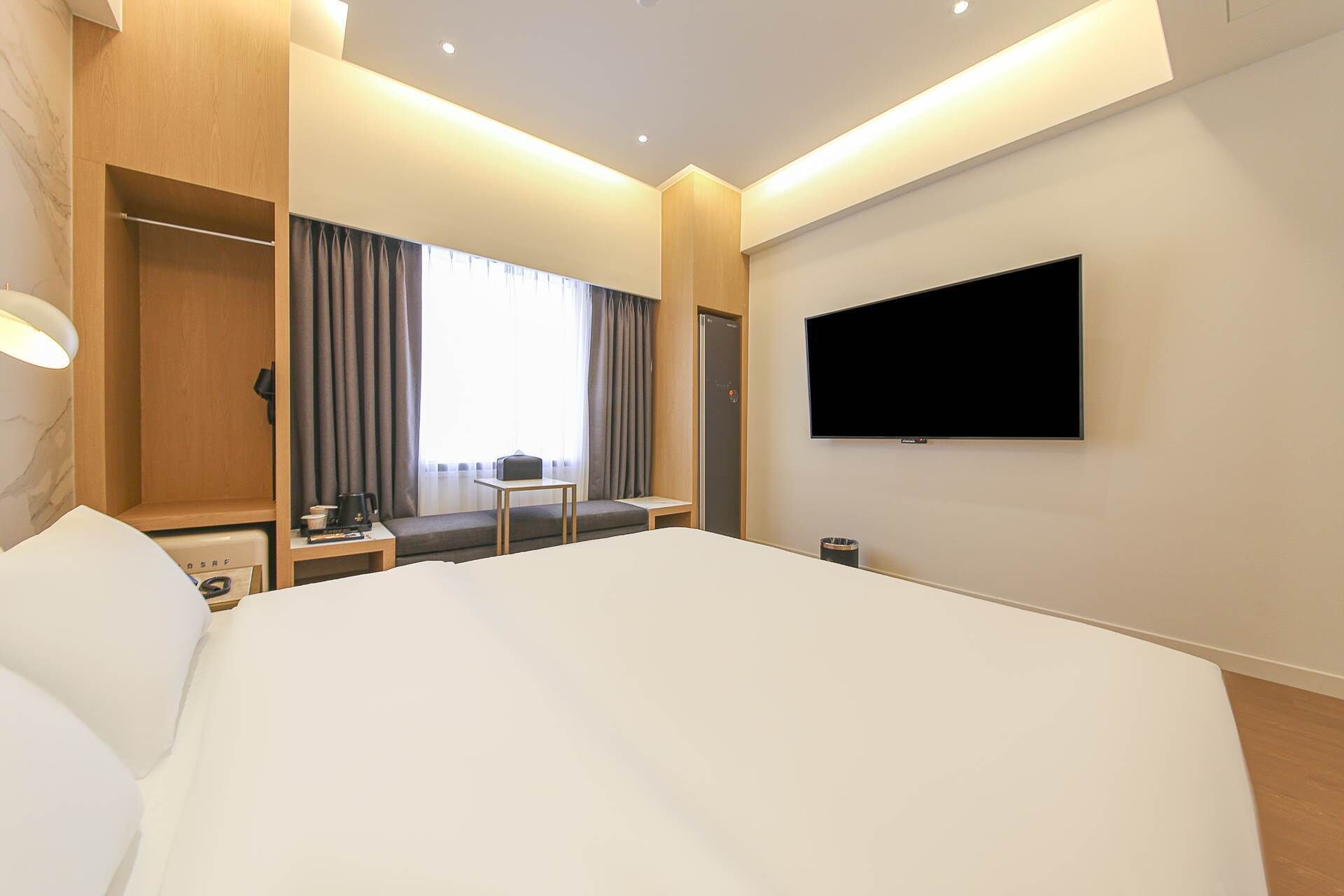 Una habitación de hotel moderna con una cama grande, una mesita de noche con lámpara, una pequeña mesa con cafetera, un televisor montado en la pared y una ventana con cortinas.