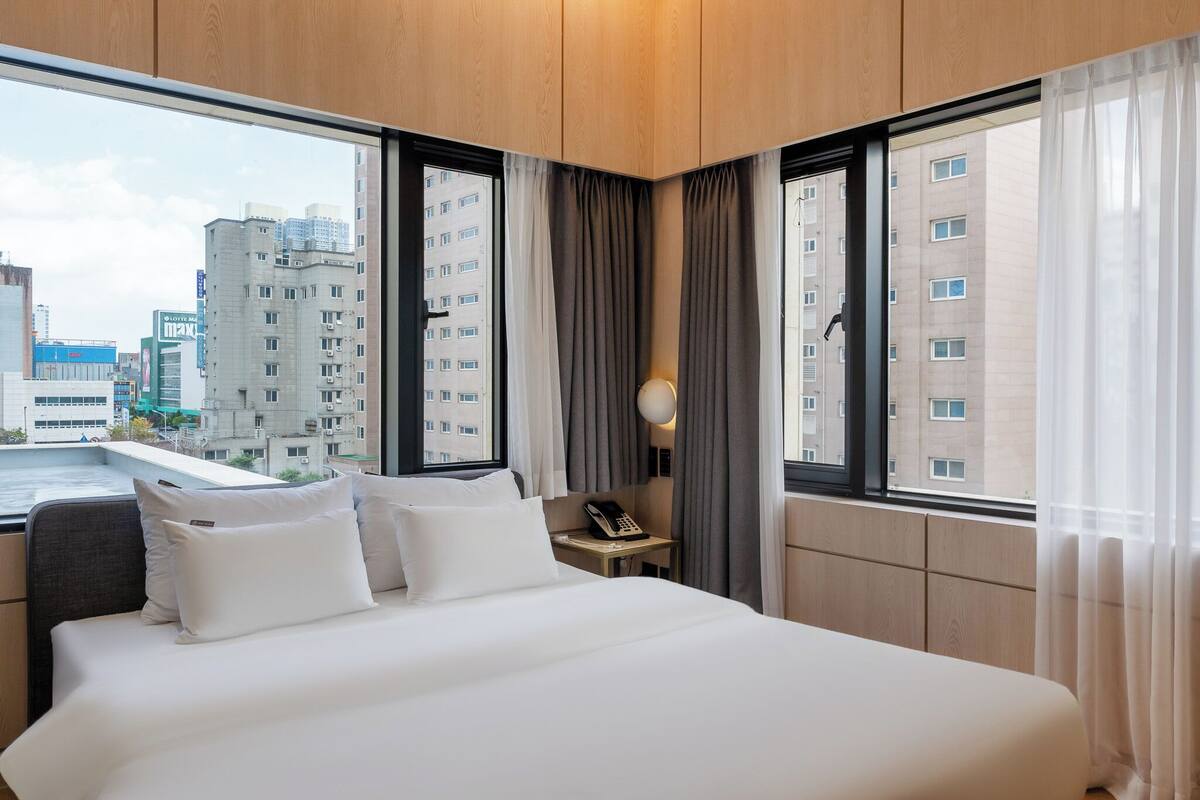 Ein Hotelzimmer mit einem großen Bett, einem Nachttisch mit Lampe und Blick auf Stadtgebäude durch große Fenster.
