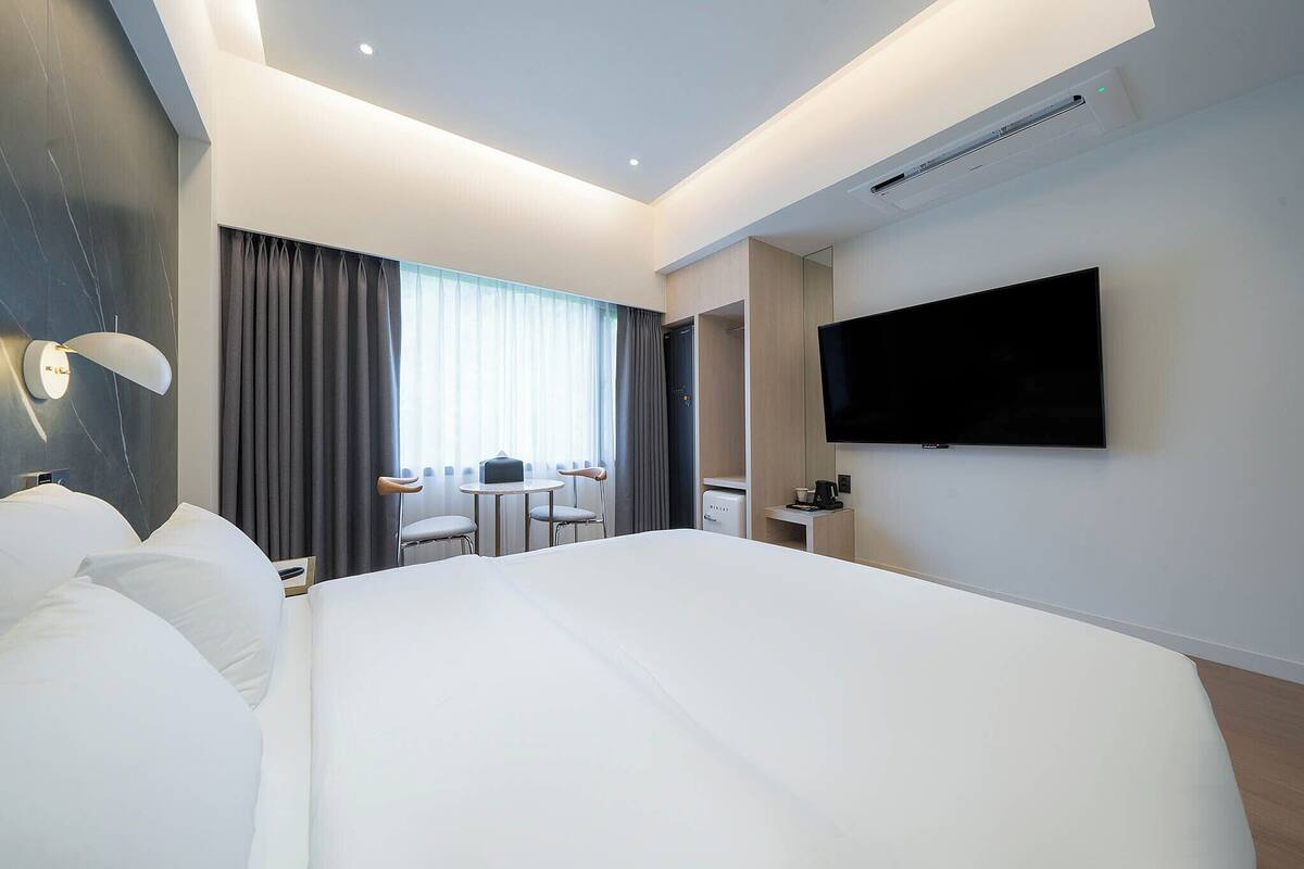 Ein modernes Hotelzimmer mit einem großen Bett, einem flachbildfernseher an der Wand, einem Schreibtisch mit Stuhl und einem kleinen Tisch mit Sitzgelegenheit.