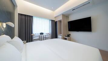 A modern hotel room with a large bed, a flat-screen TV mounted on the wall, a desk with a chair, and a small table with a chair.