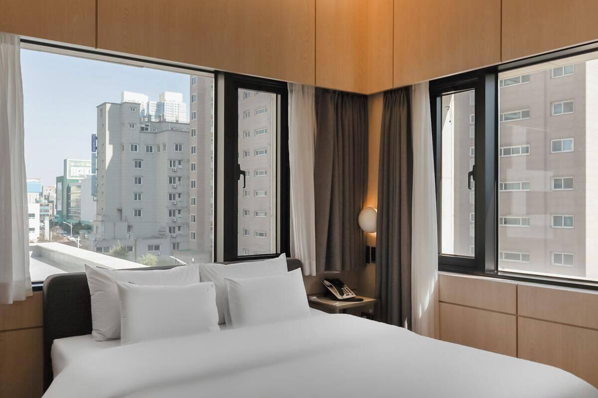 Ein Hotelzimmer mit einem großen Bett, einem Nachttisch mit Lampe und Blick auf hohe Gebäude.