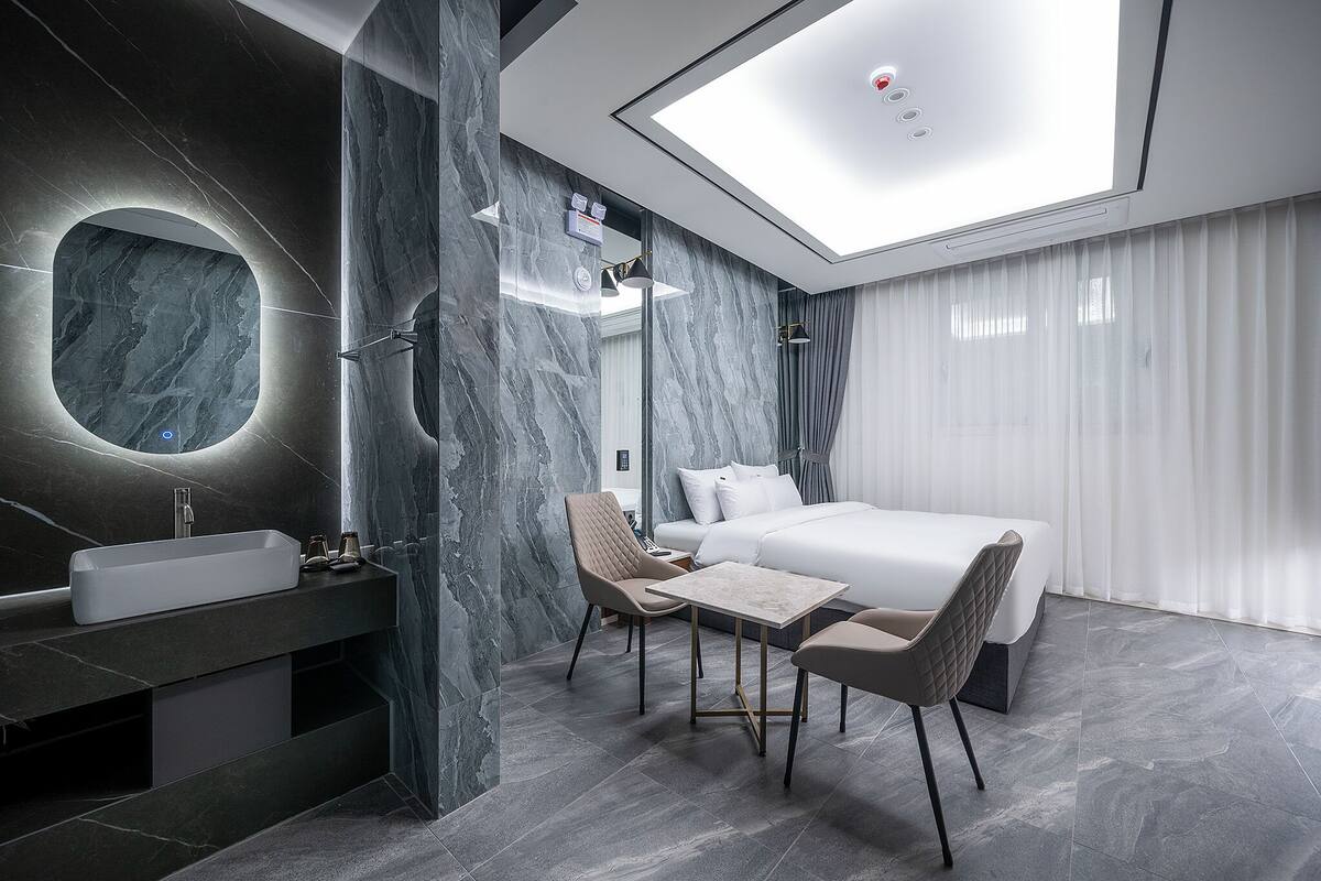Ein modernes Hotelzimmer mit einem großen Bett, einer Sitzecke mit Tisch und Stühlen, einem großen Spiegel und einem Badezimmer mit Waschbecken und Ablagefläche.