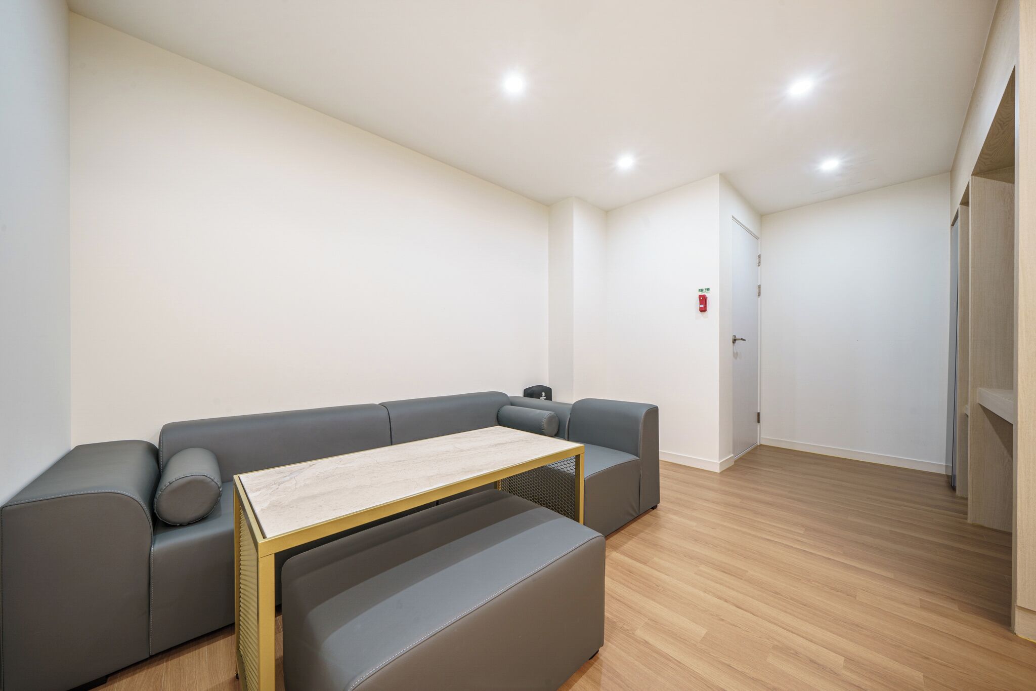A modern, minimalist room with a grey sectional sofa, a small wooden table, and a door with a fire alarm.