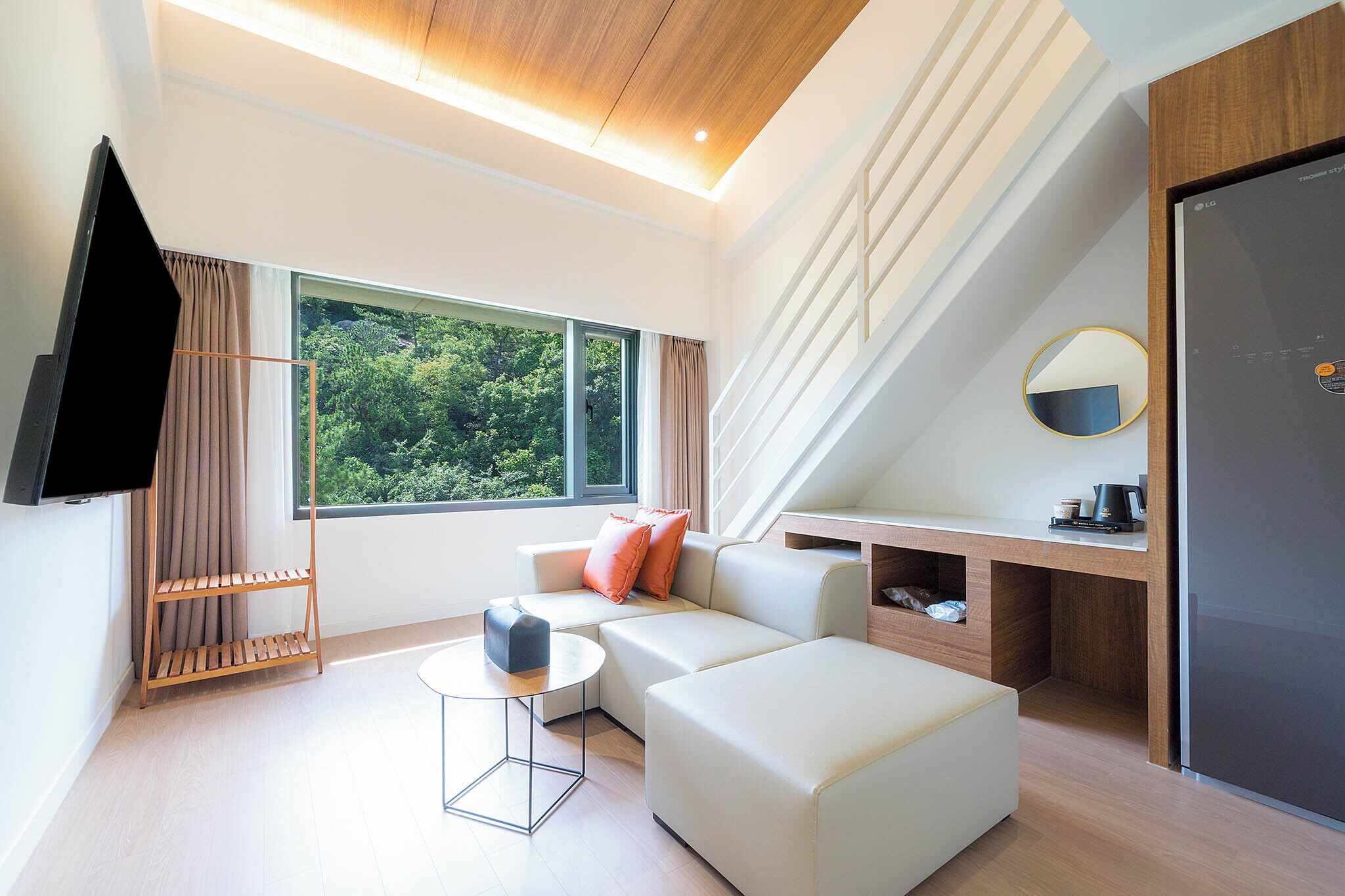 Una sala de estar moderna con un sofá, un reposapiés y una mesa de centro. Hay un televisor montado en la pared, una ventana con cortinas y un techo con vigas de madera.