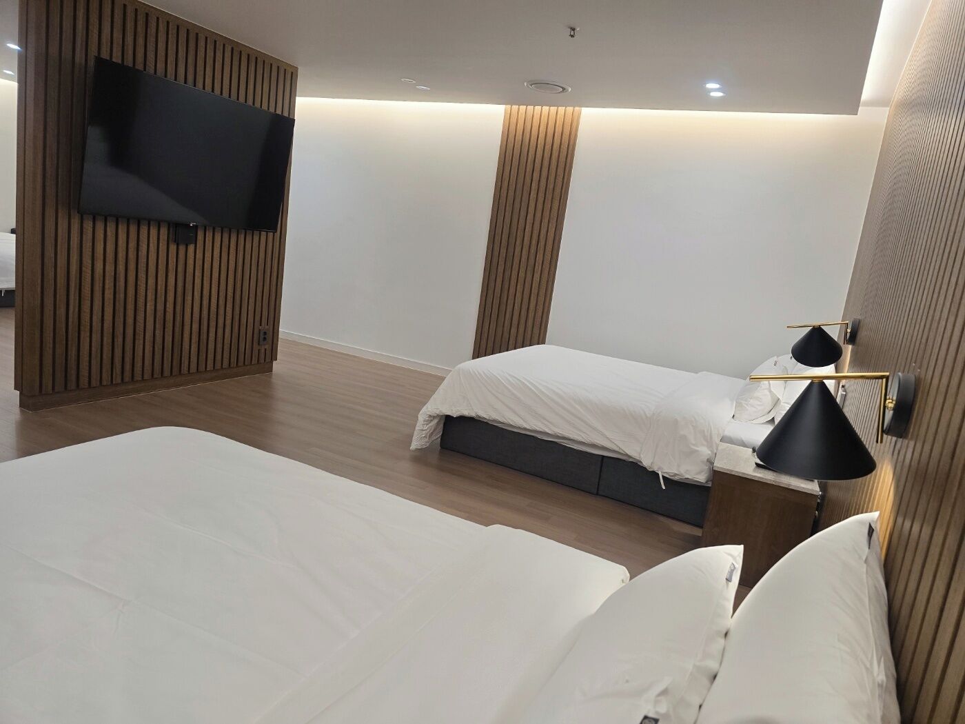 Una habitación de hotel moderna con dos camas, un televisor de pantalla plana montado en la pared y una mesita de noche con lámpara.